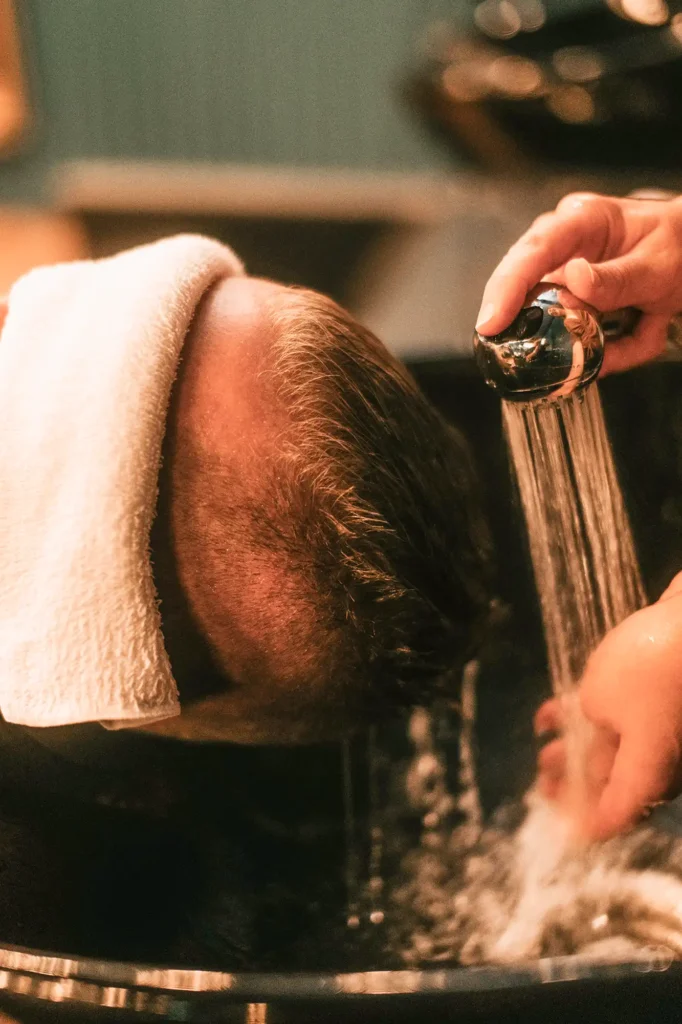 Der Kopf eines liegenden Mannes, dessen Gesicht mit einem Handtuch bedeckt ist und gerade eine Haarwäsche in einem Friseursalon bekommt.