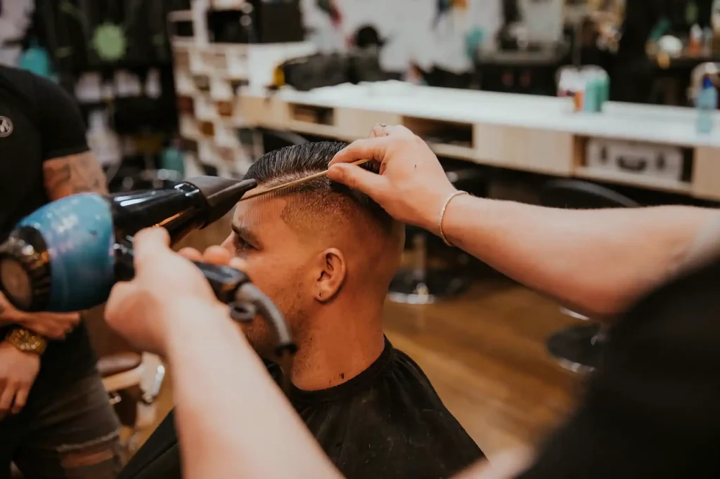 Blick auf einen Mann, der mit einem Kamm und Föhn die Haare gestyled bekommt, aus Sicht des Barbiers.